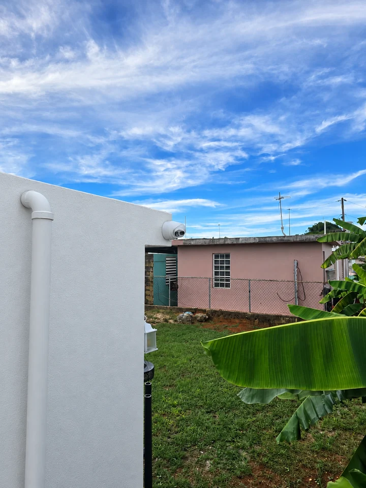 Vista amplia de una cámara sobre un muro blanco, con tubería exterior visible y vegetación al fondo.